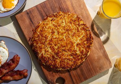 A deeply browned rÃ¶sti sits on a wooden cutting board, with plates of fried eggs and bacon, and glasses of orange juice around it on the table.