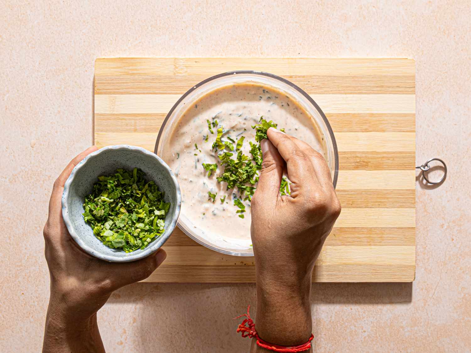 A closeup of hands garnishing a bowl of raita with fresh herbs on a wooden board