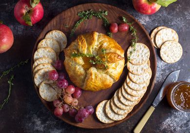 Overhead view of Baked Brie en Croûte With Thyme and Fig Jam, served on a wooden platter surrounded by grapes, shingled crackers, and herbs.