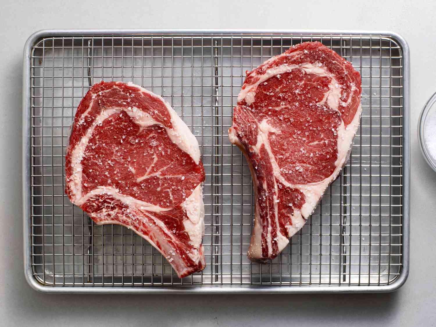 Two raw bone-in ribeyes, seasoned with kosher salt, on a wire rack set into a rimmed quarter sheet pan.