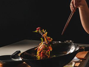 Colorful food suspended in air caused by a person tossing a wok while holding chopsticks in a dark kitchen
