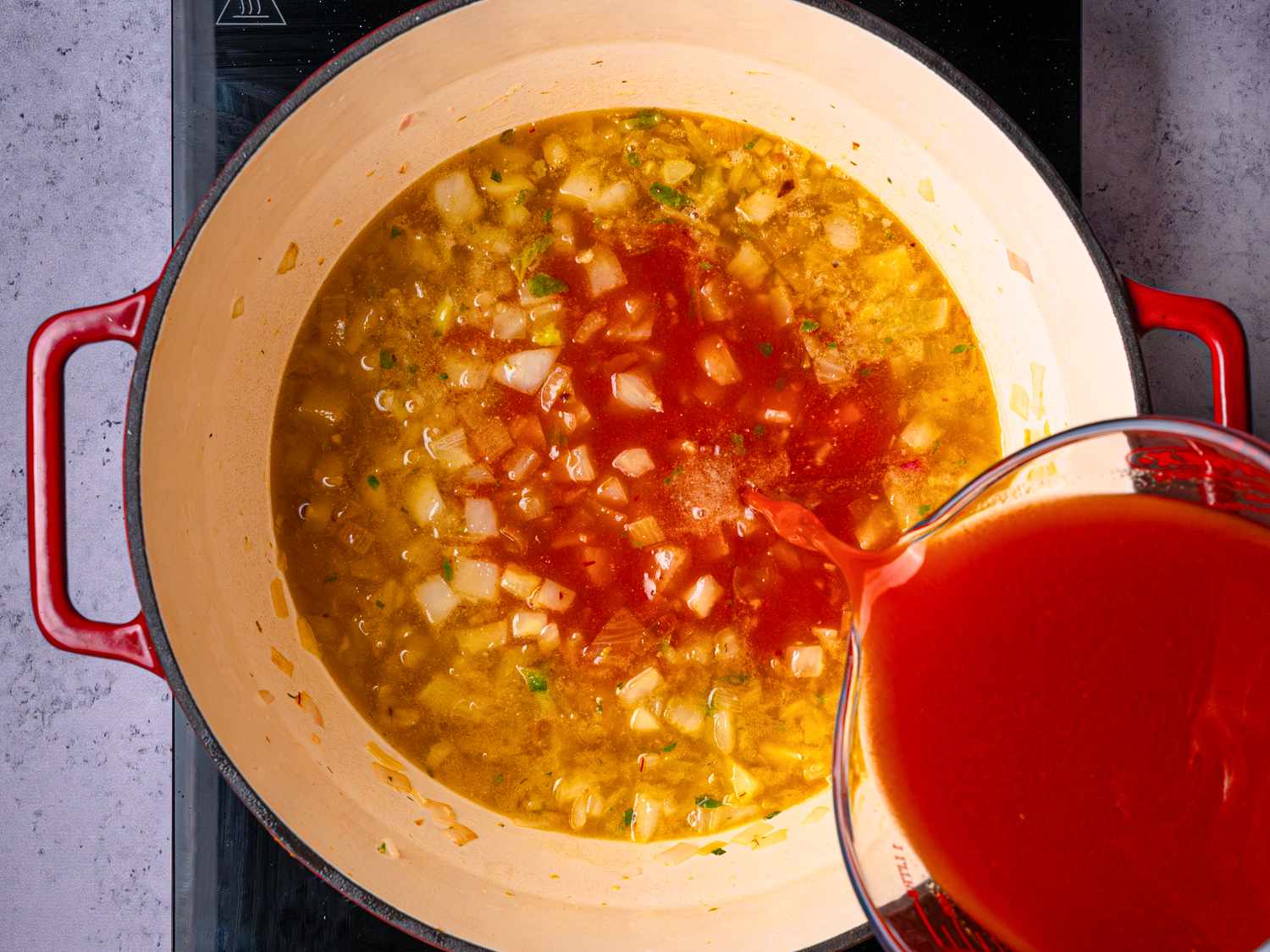 Tomato sauce being poured into a pot with diced vegetables and broth