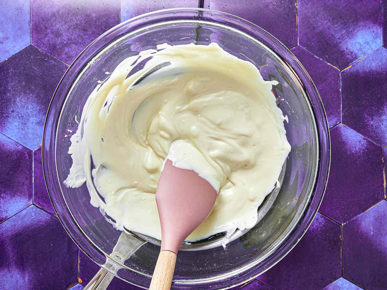 A bowl with melted white chocolate and a spatula on a purple geometric surface