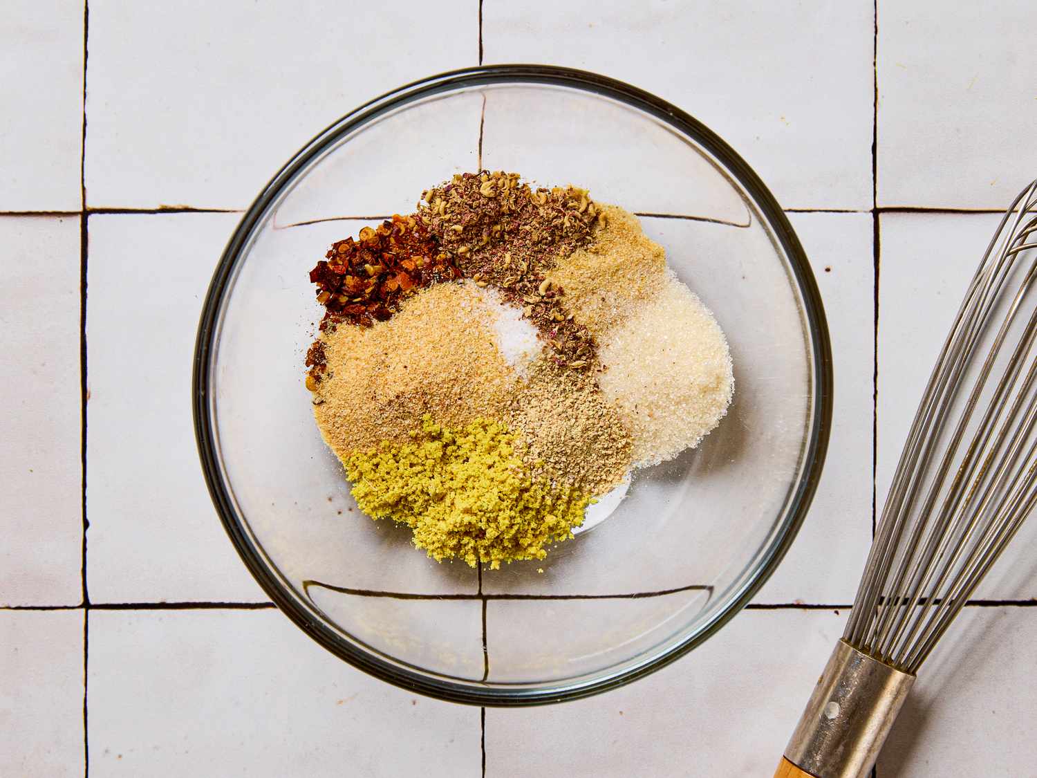 Glass bowl with various spices arranged in sections placed on a tiled surface with a whisk beside it