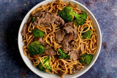 A white ceramic bowl holding lo mein with beef and broccoli on a purple colored stone surface.