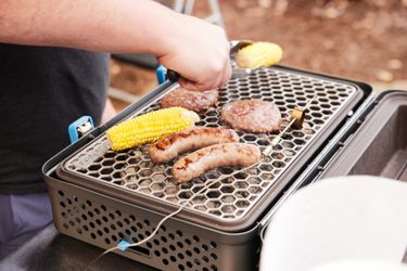 A person grilling burgers, corn, and sausages on a portable charcoal grill.