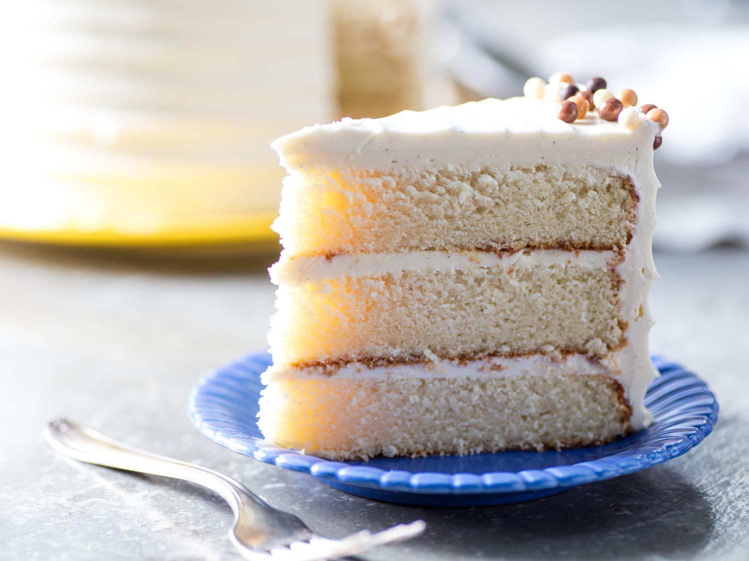 A slice of classic vanilla butter cake, served on a blue plate. The cake's crumb near the tip of the slice is diaphanous, and backlit with sunlight.
