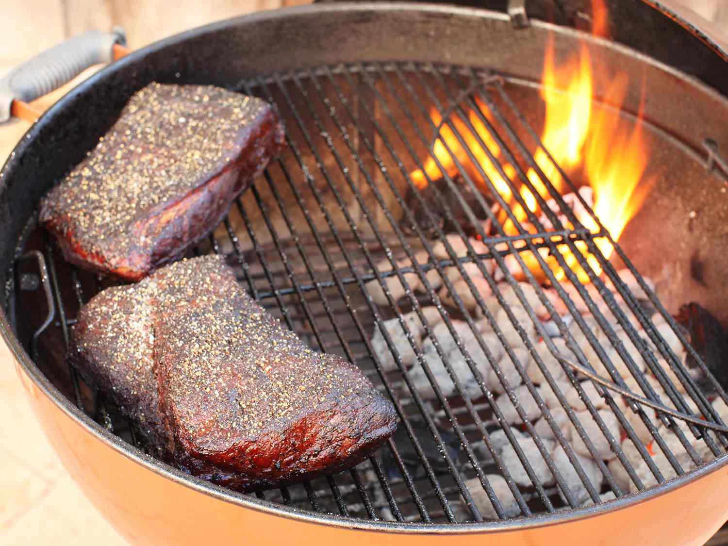 Two pieces of peppercorn-crusted brisket being smoked on small round barbecue. 