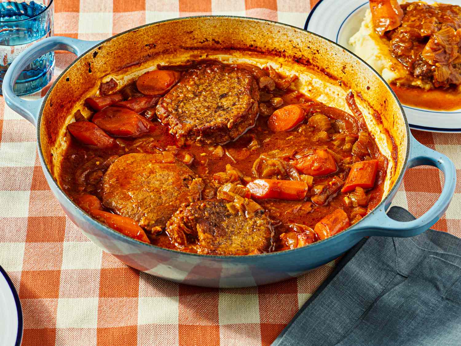 A blue casserole dish containing Swiss steak with carrots and sauce resting on a checkered tablecloth