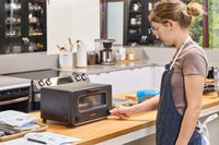 A person adjusting a knob on a balmuda toaster, situated on a countertop in a modern kitchen environment
