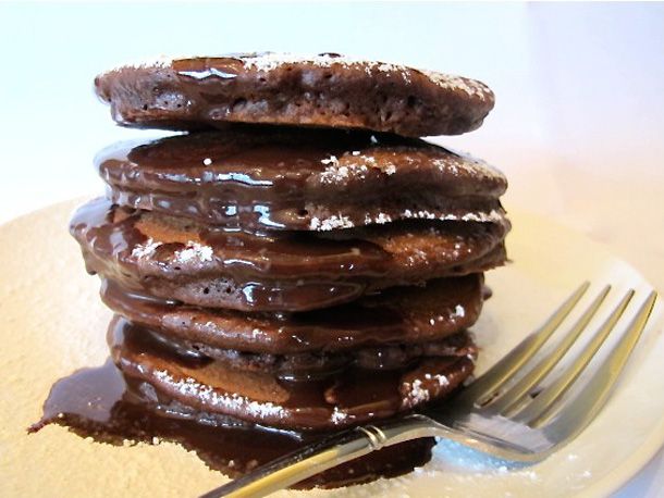 A stack of chocolate pancake on a plate, drizzled with chocolate ganache.
