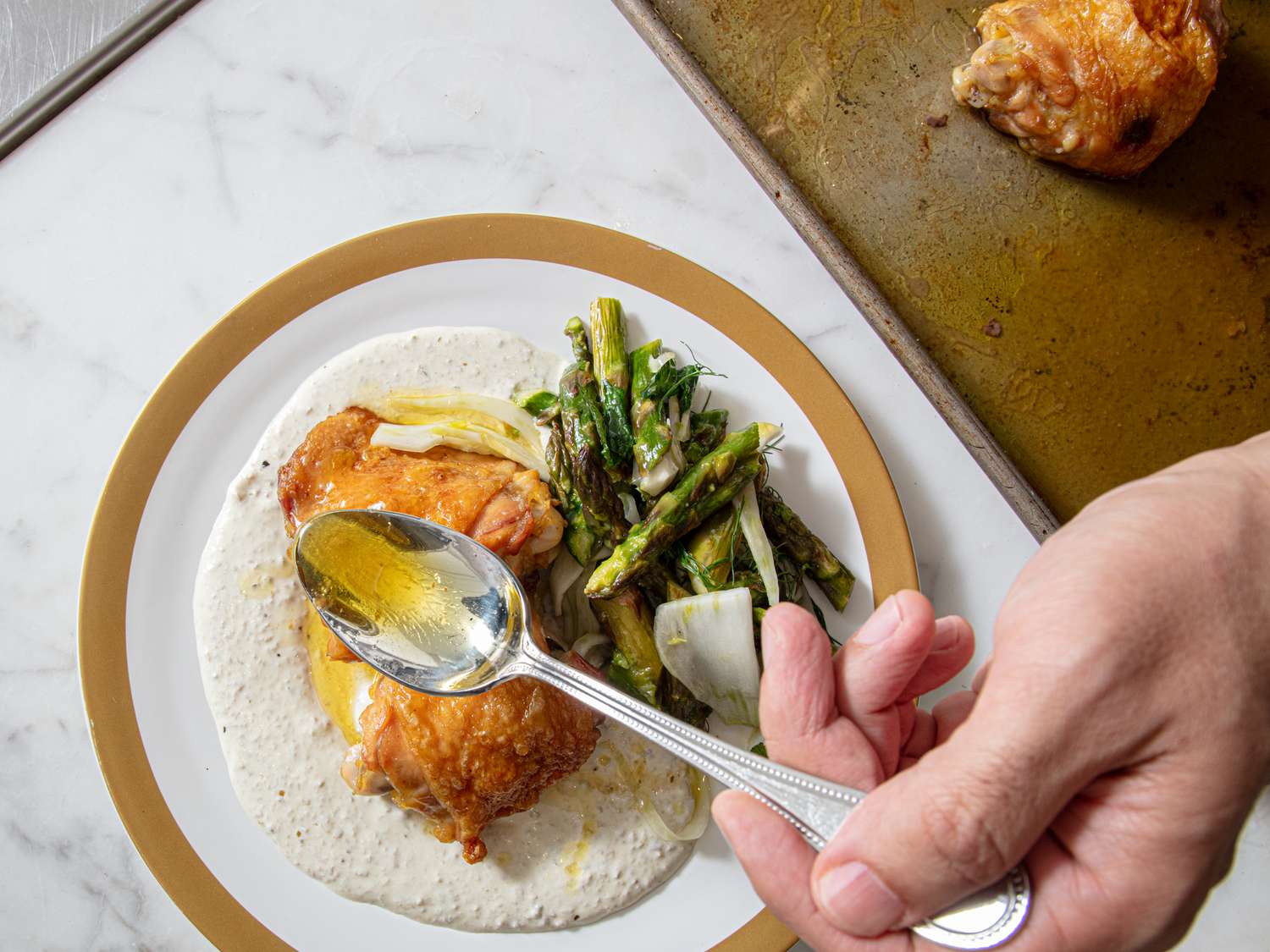 A plate with cooked chicken, vegetables, and sauce, being garnished with a spoonful of dressing or sauce