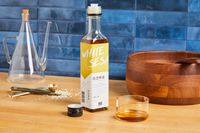 A bottle of white sesame oil on a wooden surface, accompanied by a wooden bowl, glass vessel, and measuring spoons