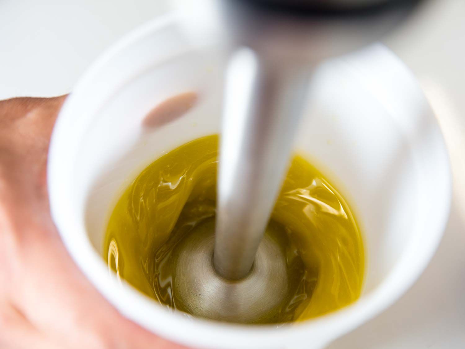 A plastic container holding olive oil being blended with an immersion blender.