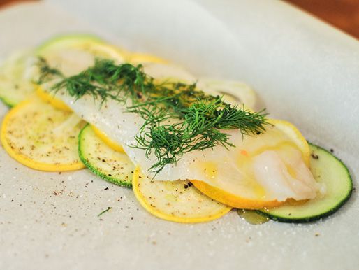 An assembled fish on parchment paper, with slices of green and yellow summer squash, lemon wheels, a fillet of fish, and fresh dill fronds.