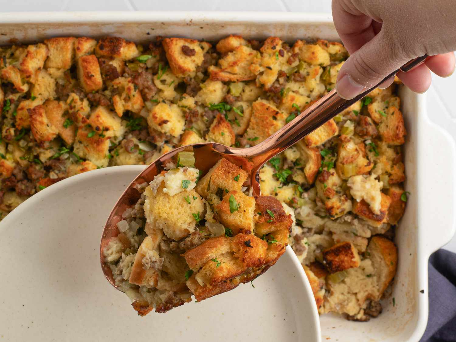 A large copper-colored serving spoon lifting a spoonful of cooked stuffing out of a pan, which is in the background.