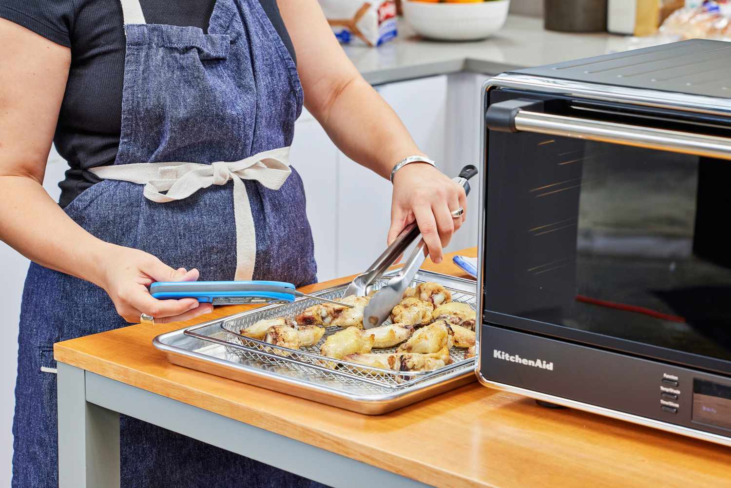 A hand tossing chicken next to the KitchenAid Dual Convection Countertop Oven with Air Fryer
