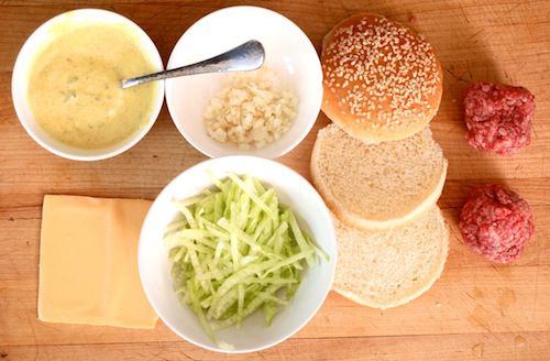Overhead view of all the ingredients needed to make a better Big Mac arranged on a wooden work surface: sauce, onion, buns sliced into three, lettuce, cheese, and two patties. 
