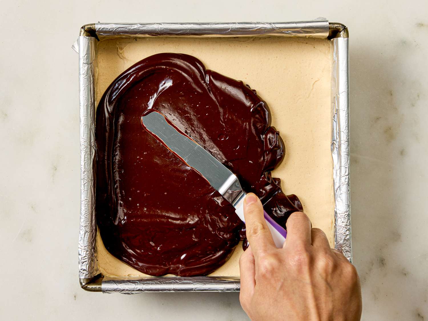 A hand spreading chocolate topping over a layered dessert in a square pan using a spatula