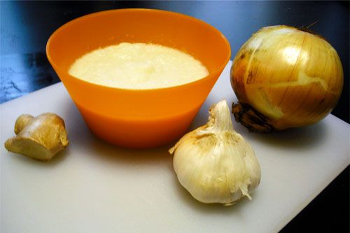 A whole onion, a bulb of garlic, and a knob of ginger are set on a cutting board next to a small bowl of onion-garlic-ginger purée.