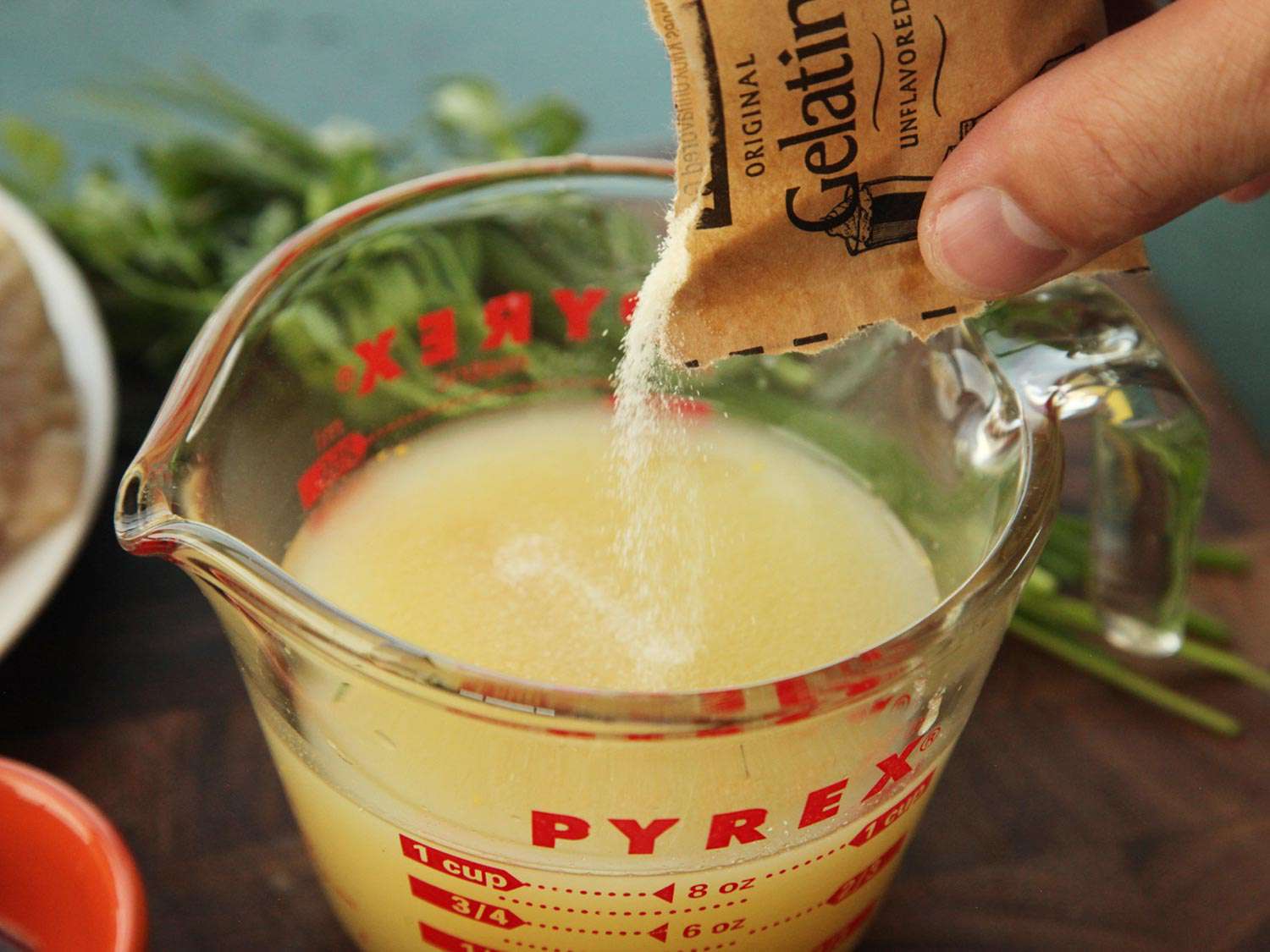 Gelatin being sprinkled over the top of a Pyrex measuring cup holding chicken stock.