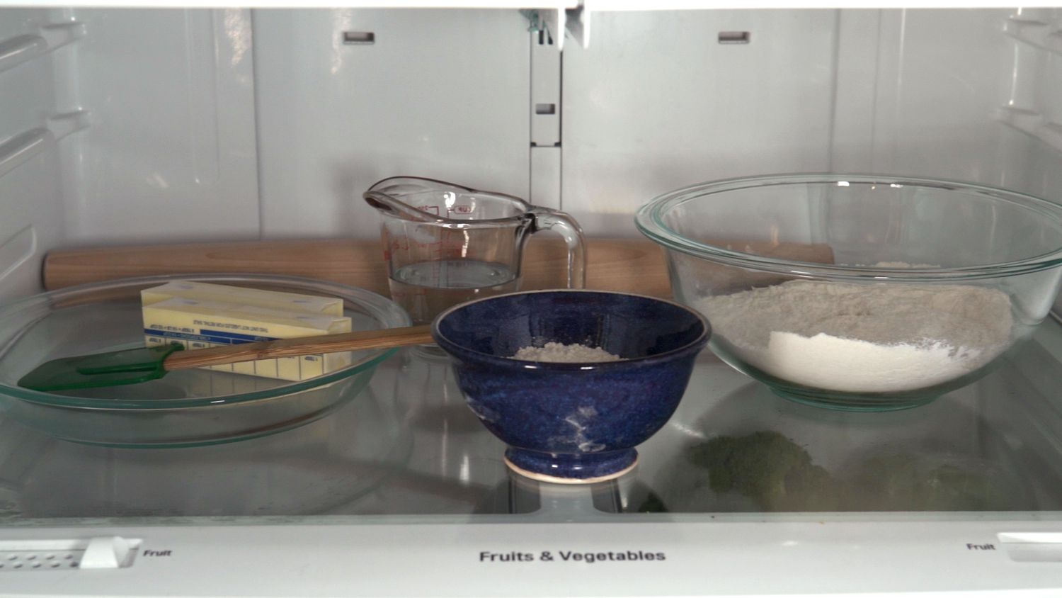 Refrigerator shelf with a glass pie dish holding a spatula and two sticks of butter, a glass measuring cup of water, a blue ceramic bowl, and a glass mixing bowl with flour