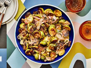Roasted Brussels sprouts and shallots with a balsamic glaze in a bowl, with glasses of sparkling water, some smaller plates and a colorfully printed table top