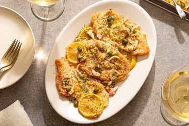 A plate of chicken piccata with lemon slices and capers on a table next to glasses of white wine