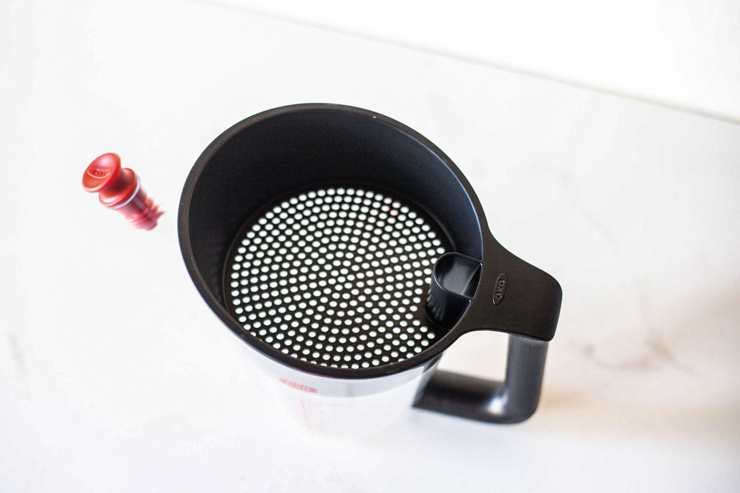 A kitchen utensil a strainer cup with a perforated lid and handle resting on a counter alongside a red stopper