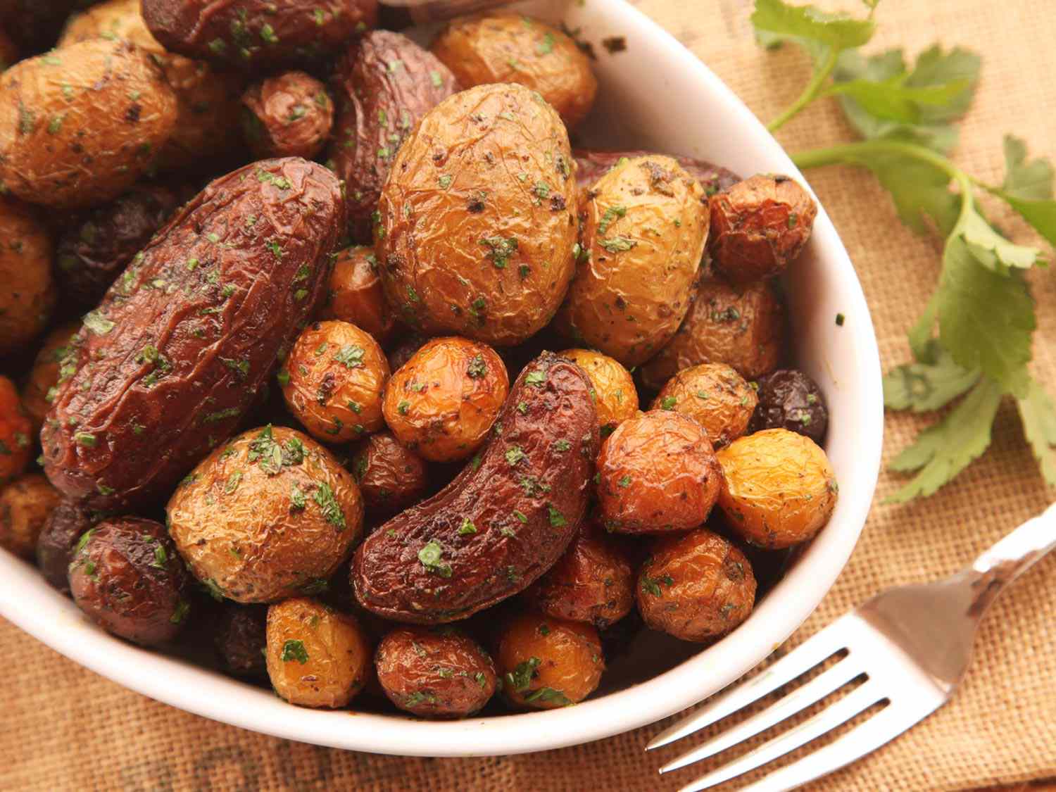 A close-up of an oval white bowl holding crispy, herb-flecked roasted potatoes.