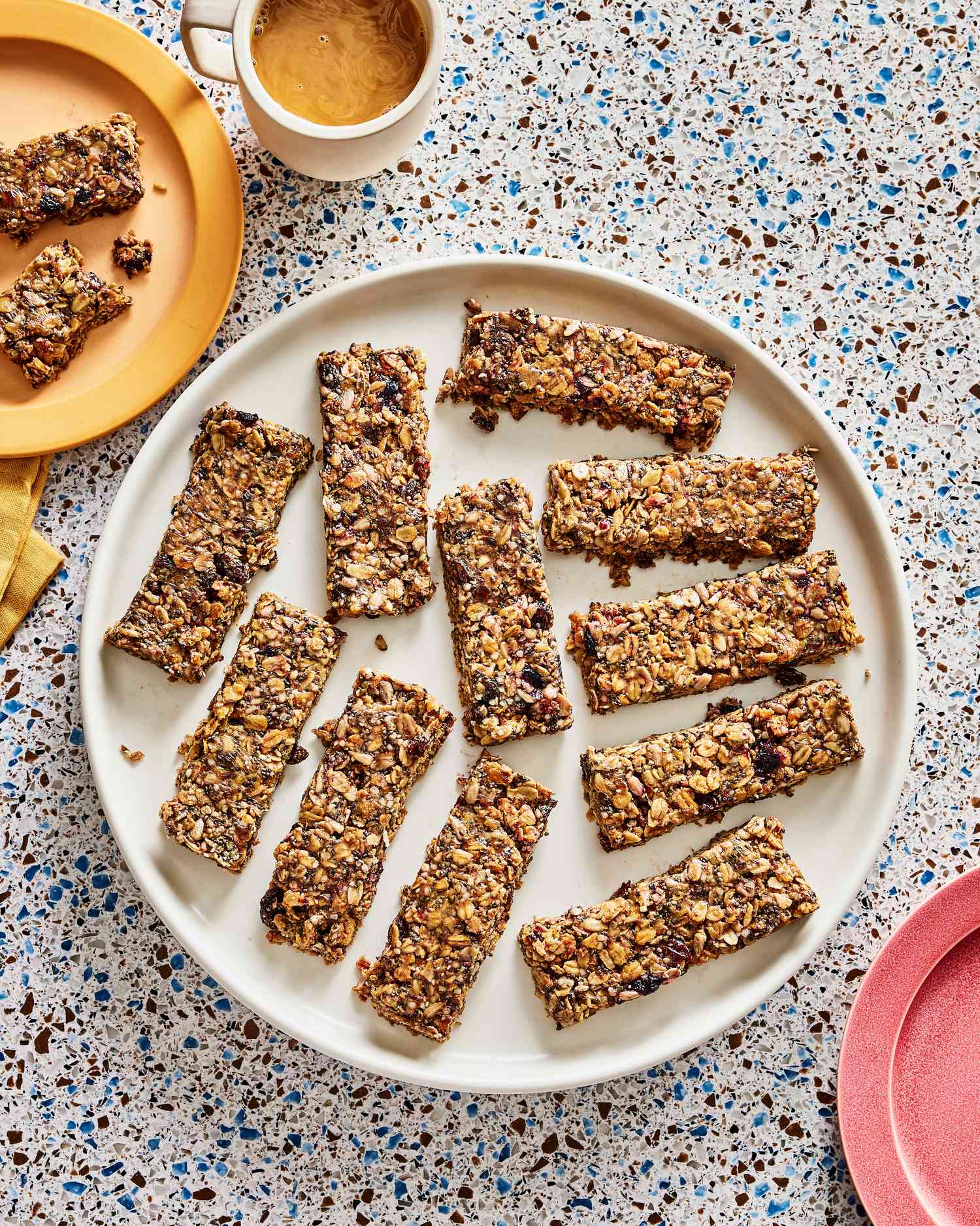 Overhead view of breakfast granola bars on a large plate. The top left has a smaller plate with a broken piece of granola bar on it, and a mug of coffee next to it. 