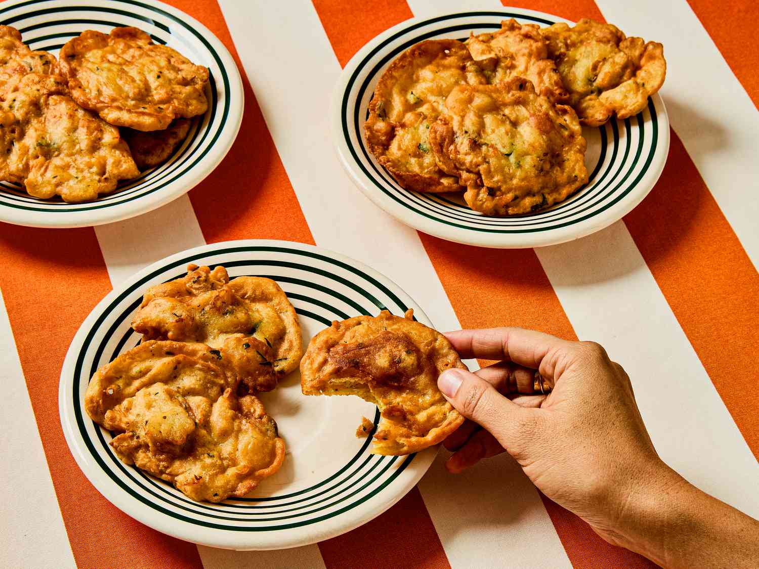 Plates of Bacalaitos on red and white striped table cloth. Hand holding a fritter with a bite taken out of it