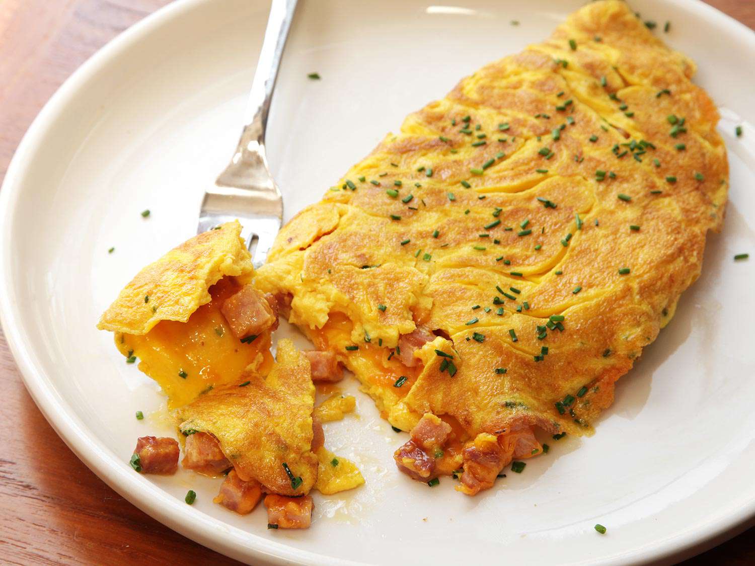 A fork removes a bite from a large ham and cheese omelette, showing off the cheesy, hammy interior.