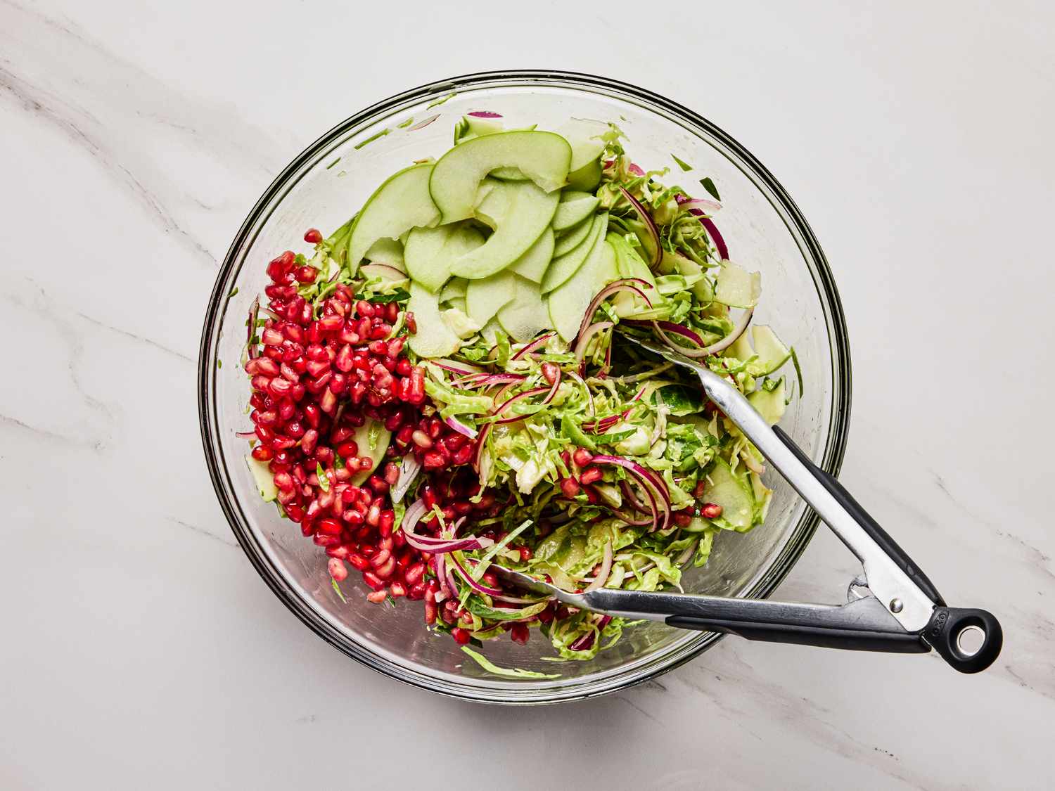 A bowl containing a salad mixture with pomegranate seeds cucumber slices and greens tongs resting on the side