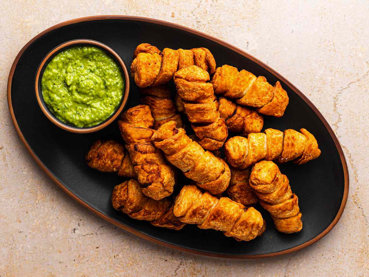 An oval black ceramic plate holding a pile of golden brown tequeños and a small bowl holding the avocado dipping sauce called guasacaca.