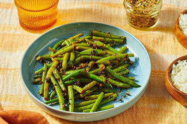 A plate of green beans seasoned with spices, next to a bowl of rice and a jar of seasonings on a textured surface