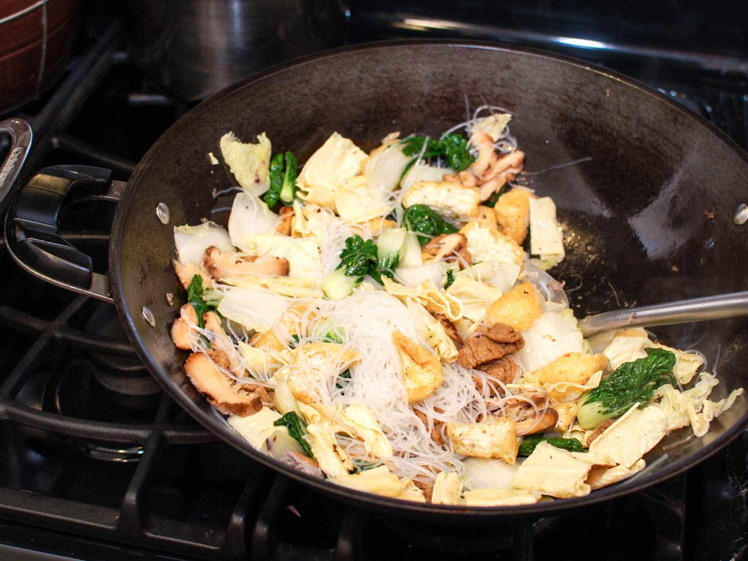 Adding vegetables to the stir-fry in the wok and stirring with a metal spatula.
