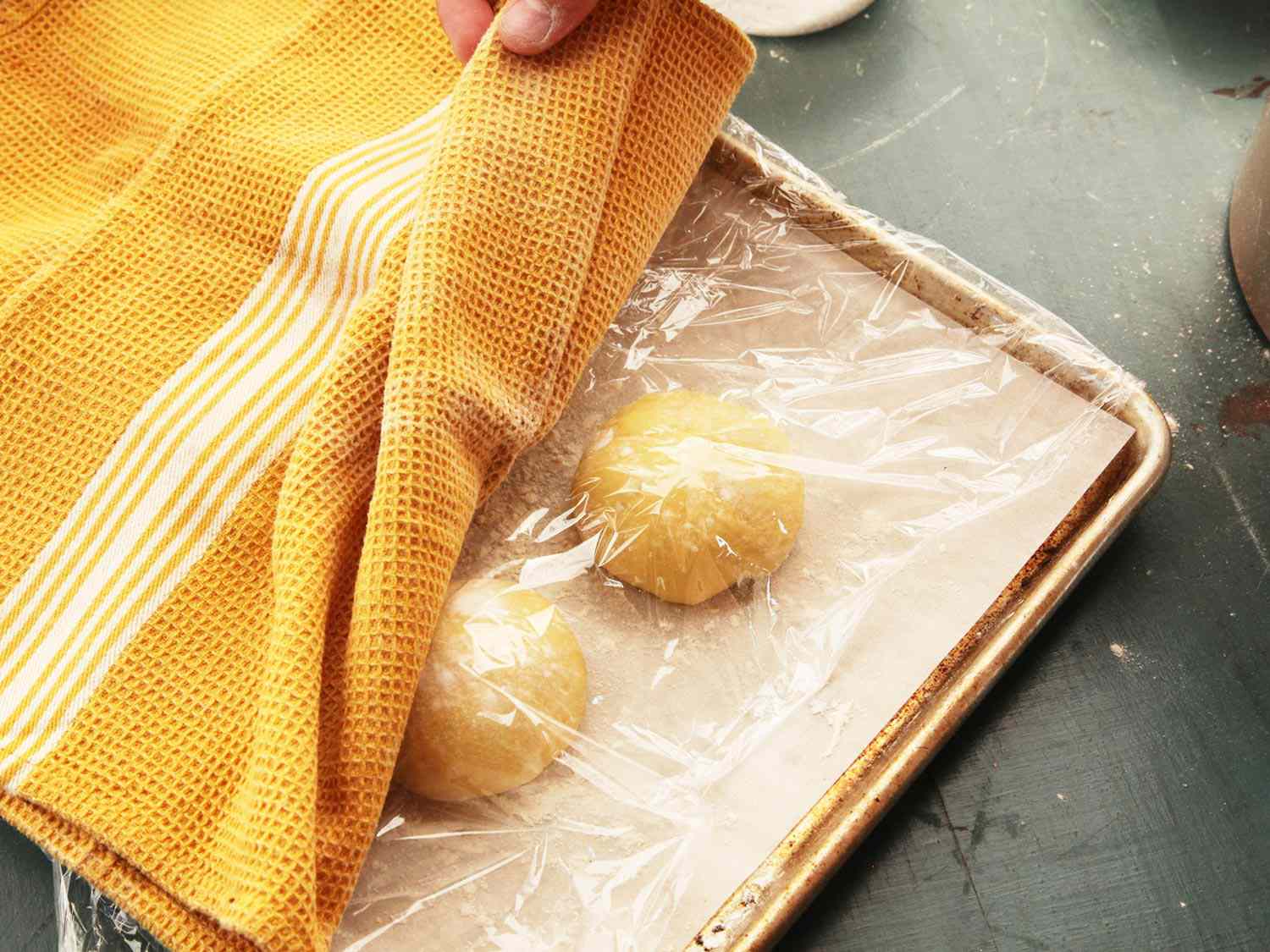Shaped cemita dough is arranged on a parchment-lined baking sheet, then covered with a layer of plastic wrap and a kitchen towel.
