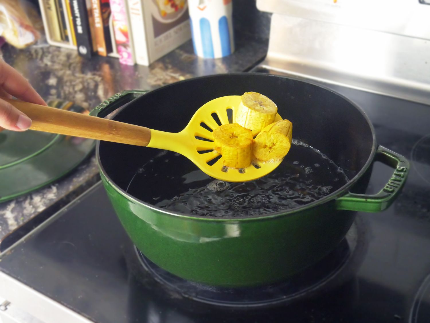 a green dutch oven with oil in it and a person using a yelloe slotted spoon to remove fried plantains