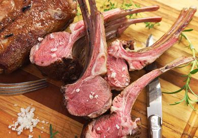 A rack of lamb cooked sous vide and sliced into individual portions, all fanned out on a wooden cutting board with a pile of salt and sprigs of fresh herbs all around.