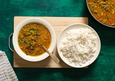 Bowl of Moong dal in a bowl, with a bowl of white rice next to it. Bowls are on a wooden board on top of green textured surface, and larger bowl of Dal is in the side of the frame. 