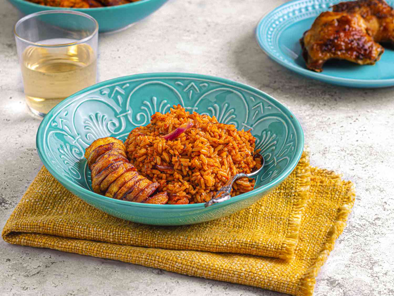 Side angle of Jollof Rice in a blue patterned bowl, on a yellow towel with chicken in the background