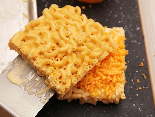 Laying a square of solid mac and cheese with a wide spatula over another square topped with shredded cheddar.