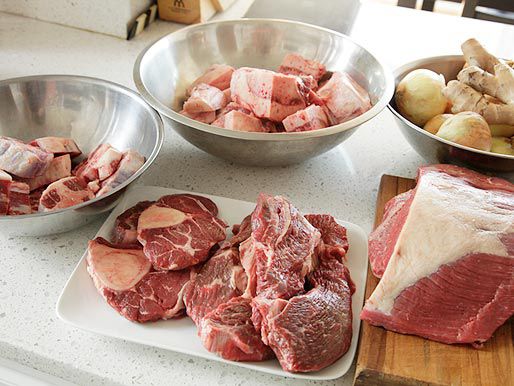 Different cuts of beef for pho: oxtail, shin, brisket, chuck, flank on the table. There's also a mixing bowl containing ginger and onion. 