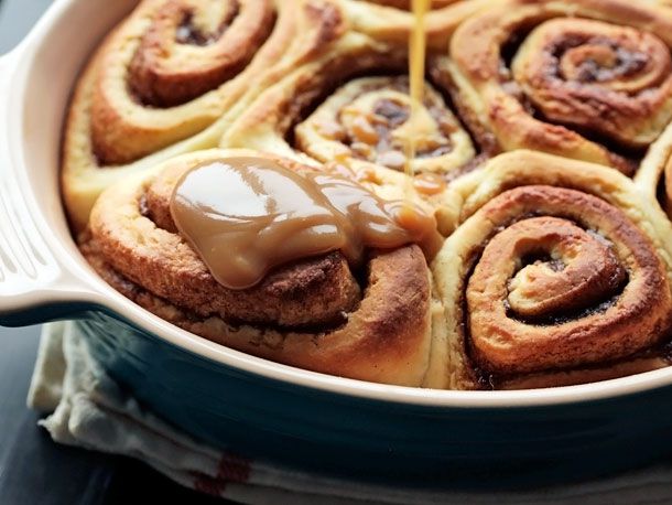 A ceramic baking dish with cinnamon sticky buns. Glaze is being drizzled over them.