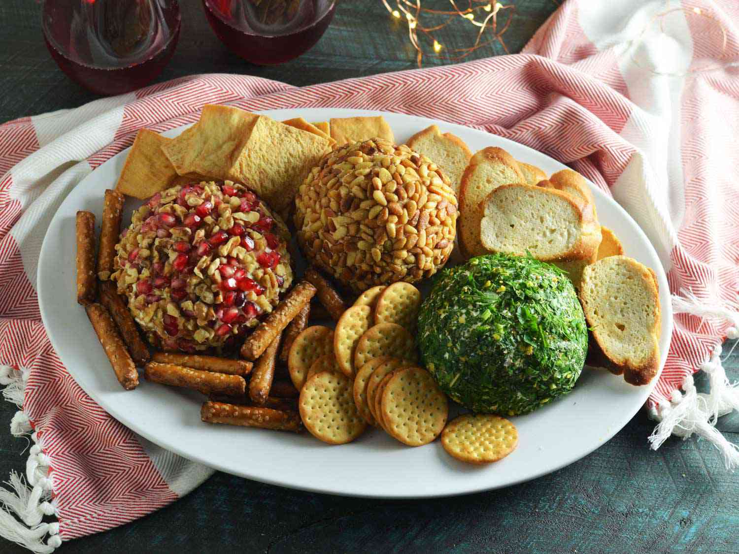 Three cheese balls on a platter, covered with different toppings. One has walnuts and pomegranate arils, the middle has toasted pine nuts, and the last is covered with a paste of green parsley and lemon zest. They are neslted on a serving platter on a bed of toasts, crackers, pretzels, and pita chips.