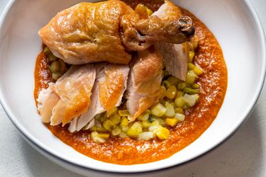 Closeup of a bowl containing sliced roast chicken pieces served atop a puddle of romesco crowned with a smattering of creamy corn.