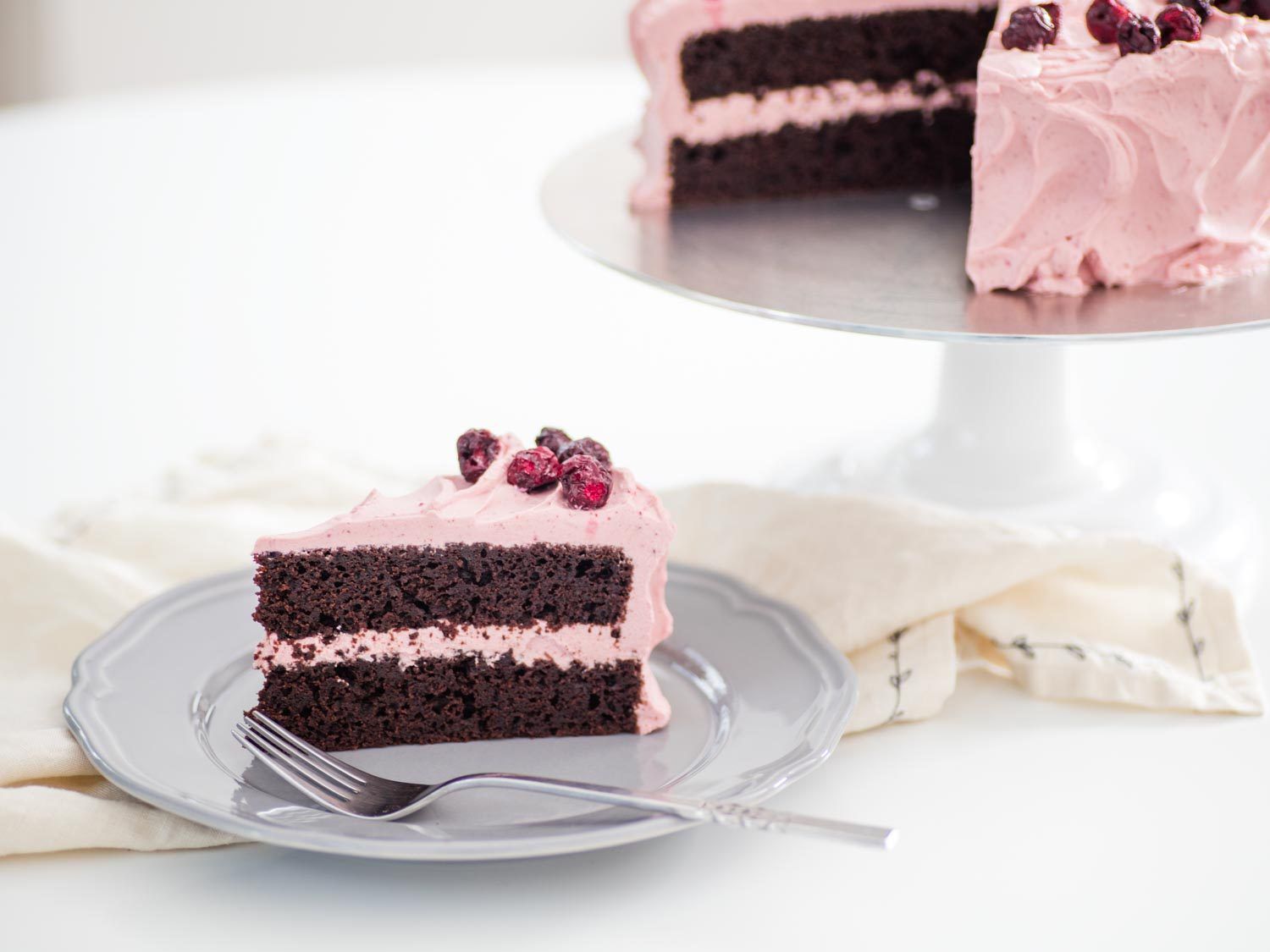 a slice of a two-layer chocolate cake with pale pink cherry whipped cream on a plate, with the remaining cake on a stand in the background
