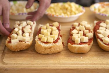 Several split french bread on a cutting board, slathered with tomato sauce and topped with differnet brands of cubed low-moisture mozzarella.