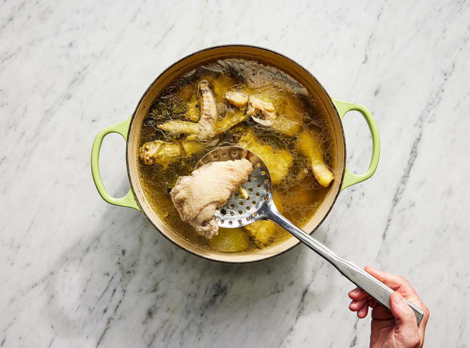 A hand lifting a piece of chicken out of the pot with a slotted spoon